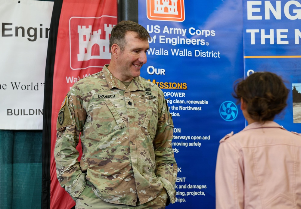 A U.S. Army soldier named Droesch, wearing Army Combat Uniform (ACU) in MultiCam pattern, is engaged in conversation with a civilian at what appears to be a recruitment or outreach event. The setting features banners for the U.S. Army Corps of Engineers, Walla Walla District, describing their missions including hydropower and waterway navigation. The soldier is smiling and appears to be in a friendly, informal discussion.