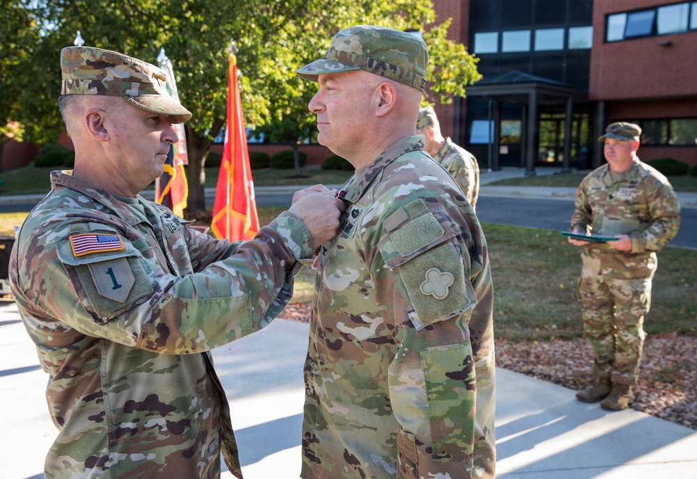 A military award or decoration ceremony is taking place outdoors at what appears to be a U.S. Army installation. A senior soldier on the left is pinning a medal or award onto the uniform of another soldier facing him. A third soldier stands in the background holding what appears to be a folder or citation.