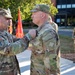 A military award or decoration ceremony is taking place outdoors at what appears to be a U.S. Army installation. A senior soldier on the left is pinning a medal or award onto the uniform of another soldier facing him. A third soldier stands in the background holding what appears to be a folder or citation.