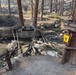 A picnic area in a forested campground shows significant wildfire damage. A wooden footbridge over a small creek has been destroyed, leaving only charred debris and burned structural remnants in the water. The surrounding trees and ground show evidence of recent fire passage, with blackened bark and scorched undergrowth.