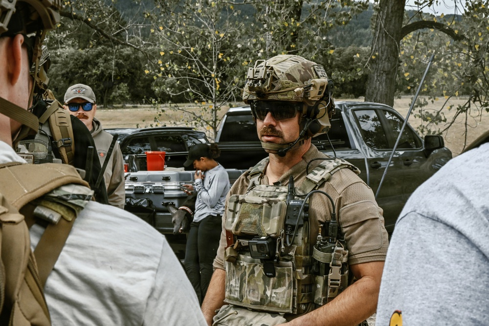 A tactical operator in full multicam gear, helmet with NVG mount, and communications headset appears to be briefing or debriefing a small group in an outdoor rural setting. Several other individuals are present, including one in civilian clothes working near the tailgate of a dark pickup truck with equipment cases. The setting suggests a field training exercise or operational staging area.