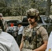 A tactical operator in full multicam gear, helmet with NVG mount, and communications headset appears to be briefing or debriefing a small group in an outdoor rural setting. Several other individuals are present, including one in civilian clothes working near the tailgate of a dark pickup truck with equipment cases. The setting suggests a field training exercise or operational staging area.
