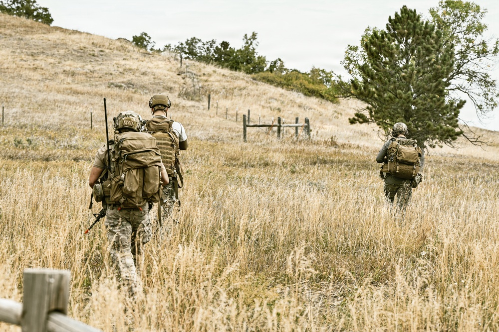 Three armed and equipped military or tactical personnel are moving through a dry grassland field toward a hillside. They are viewed from behind, wearing full tactical gear including helmets, body armor, and large rucksacks. The setting appears to be a rural training area or operational environment with rolling hills and sparse trees.