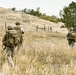 Three armed and equipped military or tactical personnel are moving through a dry grassland field toward a hillside. They are viewed from behind, wearing full tactical gear including helmets, body armor, and large rucksacks. The setting appears to be a rural training area or operational environment with rolling hills and sparse trees.