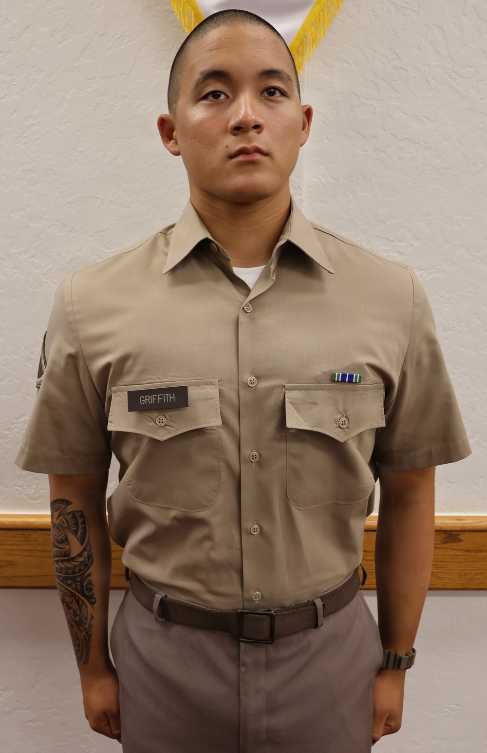 A young male service member in a khaki military uniform stands at attention in front of a white textured wall with a partially visible flag or pennant with gold fringe in the background. He wears a name tape reading 'GRIFFITH' on his left chest pocket and a ribbon rack on his right chest. He has a visible tattoo on his left forearm.