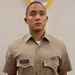 A young male service member in a khaki military uniform stands at attention in front of a white textured wall with a partially visible flag or pennant with gold fringe in the background. He wears a name tape reading 'GRIFFITH' on his left chest pocket and a ribbon rack on his right chest. He has a visible tattoo on his left forearm.