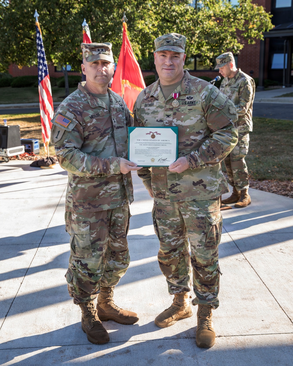 Two U.S. Army soldiers pose together holding a Meritorious Service Medal certificate during an outdoor awards ceremony. The soldier on the left, identified by name tape as 'MEISLER,' wears two-star Major General rank insignia, while the soldier on the right, identified as 'OSTHOFF,' wears Colonel (full bird) rank and has a medal pinned to his chest. A third soldier is visible in the background near a microphone stand.