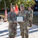 Two U.S. Army soldiers pose together holding a Meritorious Service Medal certificate during an outdoor awards ceremony. The soldier on the left, identified by name tape as 'MEISLER,' wears two-star Major General rank insignia, while the soldier on the right, identified as 'OSTHOFF,' wears Colonel (full bird) rank and has a medal pinned to his chest. A third soldier is visible in the background near a microphone stand.