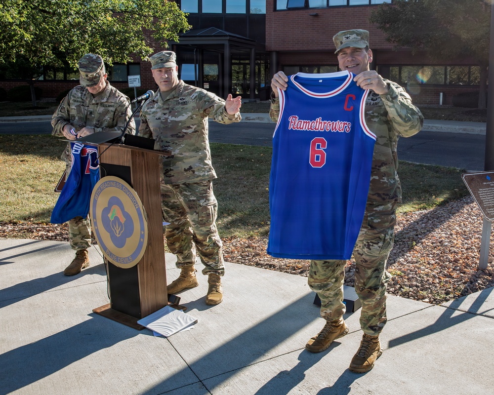 Three U.S. Army soldiers in camouflage uniforms are gathered outdoors at a podium bearing a military unit seal. One soldier is prominently displaying a blue basketball jersey reading 'Flamethrowers' with the number 6 and a 'C' (captain) designation. The setting appears to be a formal outdoor ceremony or presentation at a military installation.