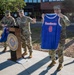 Three U.S. Army soldiers in camouflage uniforms are gathered outdoors at a podium bearing a military unit seal. One soldier is prominently displaying a blue basketball jersey reading 'Flamethrowers' with the number 6 and a 'C' (captain) designation. The setting appears to be a formal outdoor ceremony or presentation at a military installation.