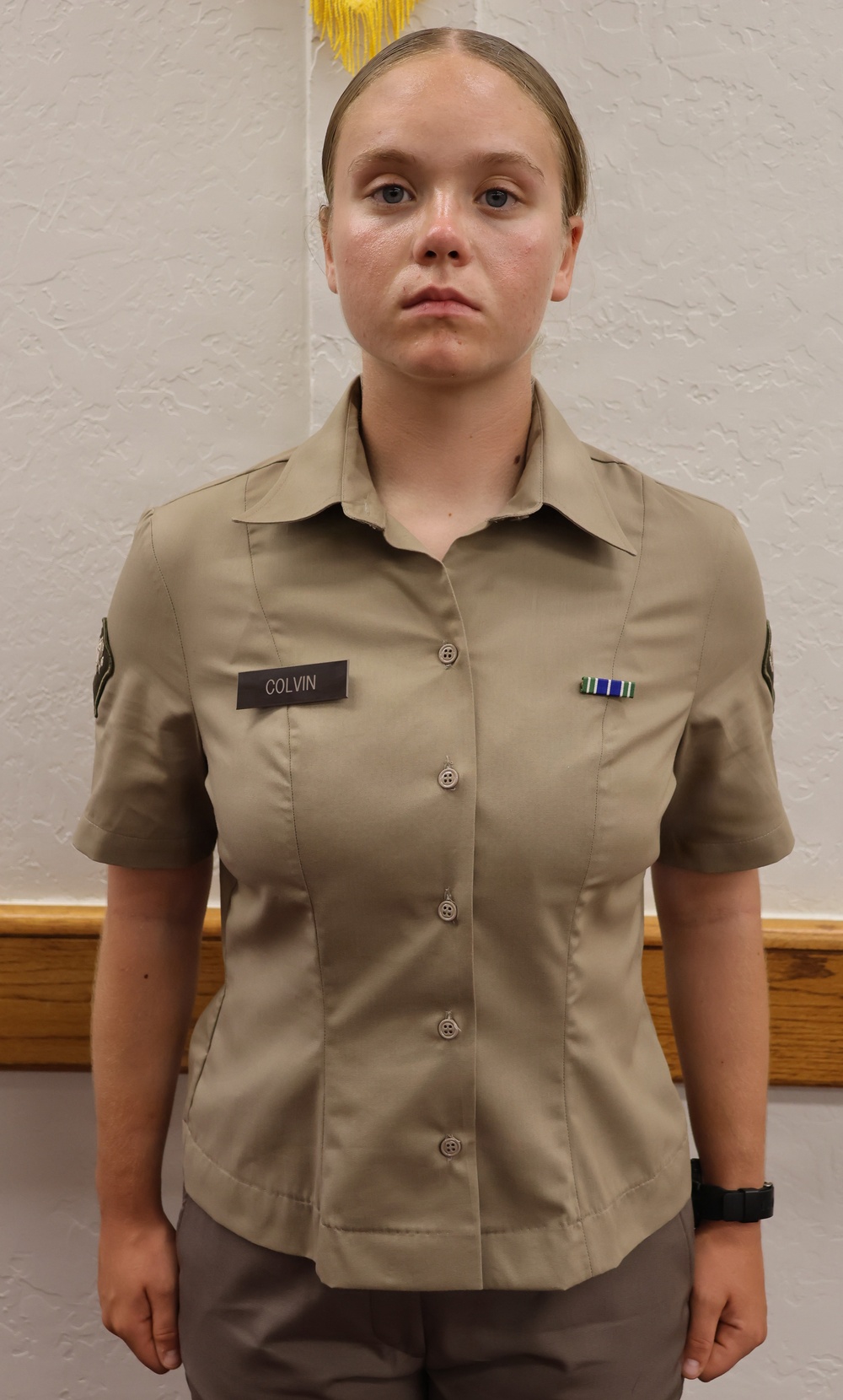 A young person in a khaki military-style uniform stands at attention against a white textured wall. The uniform displays a name tape reading 'COLVIN', a ribbon medal on the right chest, and a patch on the left sleeve. The setting appears to be an official or formal military/JROTC portrait.