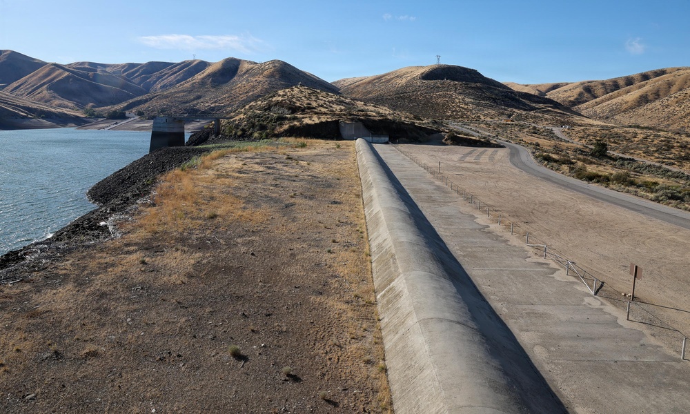 The image shows a large earthen dam with a concrete spillway or parapet wall running along its crest, adjacent to a reservoir with notably low water levels. The surrounding landscape consists of dry, brown hills characteristic of an arid or semi-arid region, suggesting drought conditions. Infrastructure elements including a concrete intake/outlet structure and power transmission lines are visible in the background.