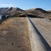 The image shows a large earthen dam with a concrete spillway or parapet wall running along its crest, adjacent to a reservoir with notably low water levels. The surrounding landscape consists of dry, brown hills characteristic of an arid or semi-arid region, suggesting drought conditions. Infrastructure elements including a concrete intake/outlet structure and power transmission lines are visible in the background.