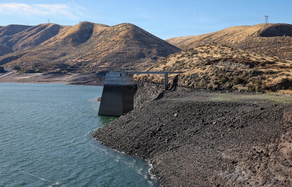 The image shows a reservoir at significantly low water levels, exposing a large expanse of dark rocky shoreline. A concrete intake tower or control structure stands near the water's edge, connected to the shore by a bridge or walkway. Arid, golden-brown hills with power transmission lines frame the background, suggesting a drought-affected water storage facility in a semi-arid region.