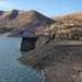 The image shows a reservoir at significantly low water levels, exposing a large expanse of dark rocky shoreline. A concrete intake tower or control structure stands near the water's edge, connected to the shore by a bridge or walkway. Arid, golden-brown hills with power transmission lines frame the background, suggesting a drought-affected water storage facility in a semi-arid region.