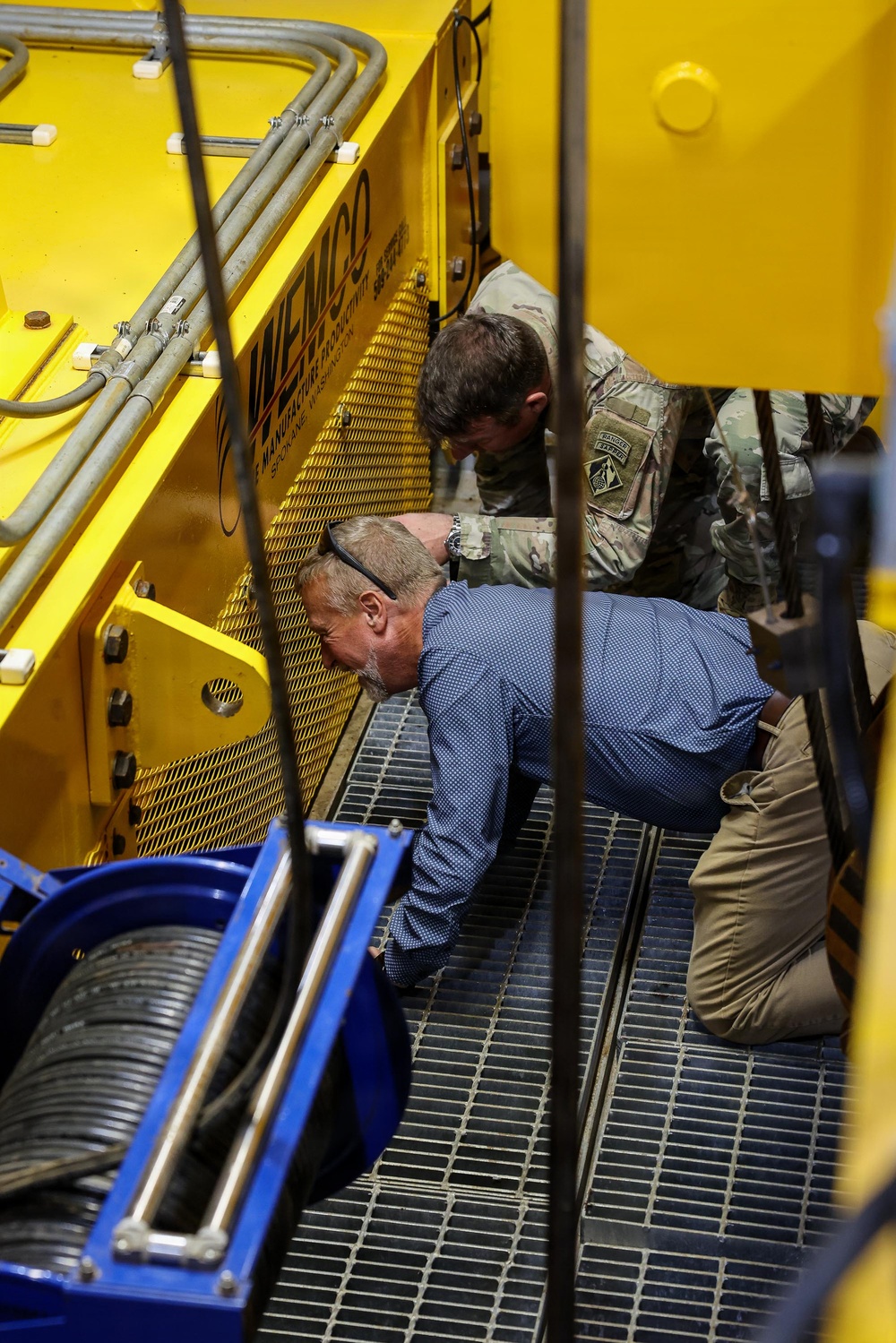 Two individuals are crouched down inspecting the underside or interior components of a large yellow industrial machine manufactured by WEMCO, based in Spokane, Washington. One person is wearing military camouflage uniform with visible patches including what appears to be a Ranger tab, while the other is in civilian business casual attire. They appear to be conducting an inspection or technical review of the equipment.