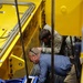 Two individuals are crouched down inspecting the underside or interior components of a large yellow industrial machine manufactured by WEMCO, based in Spokane, Washington. One person is wearing military camouflage uniform with visible patches including what appears to be a Ranger tab, while the other is in civilian business casual attire. They appear to be conducting an inspection or technical review of the equipment.
