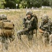 Three heavily equipped tactical personnel are positioned in a field of tall dry grass, wearing full combat gear including helmets, plate carriers, and tactical backpacks. The central figure is standing and scanning the area while the right figure appears to be kneeling and operating a device. The setting appears to be a rural or semi-rural area with trees and a white fence visible in the background.