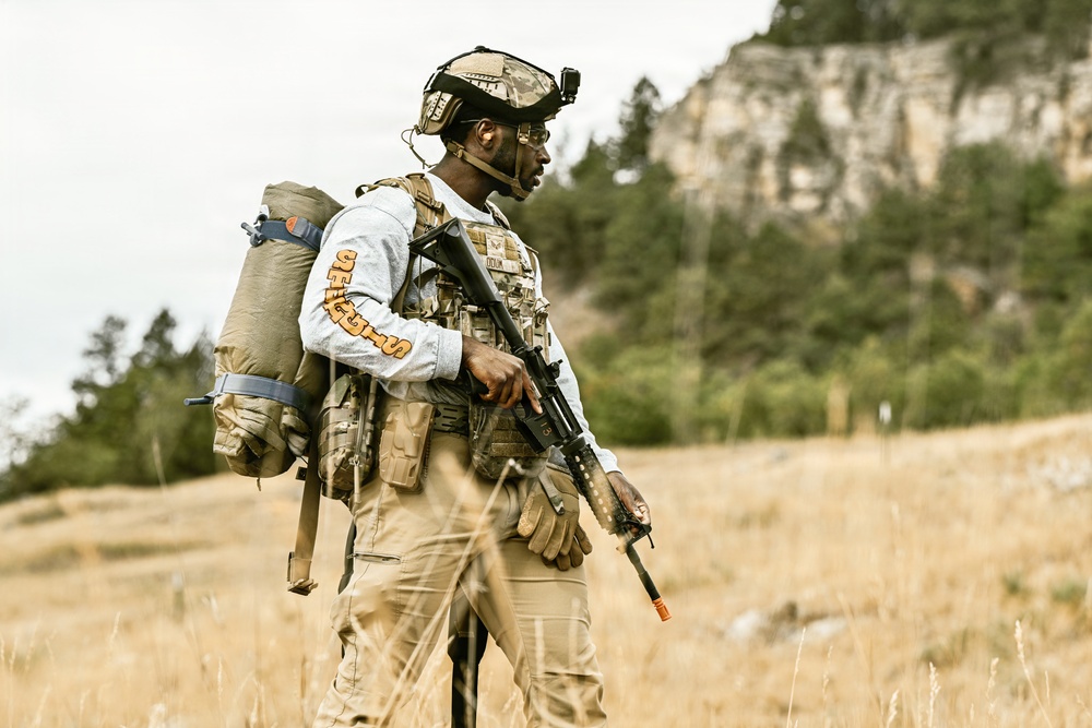 A man in tactical military-style gear stands in an open field with dry grass, carrying a rifle and a large backpack. He is wearing a camouflage plate carrier vest, tactical helmet with mounted device, and gloves, and appears to be scanning his surroundings. The background features rocky cliffs, conifer trees, and an overcast sky.
