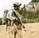 A man in tactical military-style gear stands in an open field with dry grass, carrying a rifle and a large backpack. He is wearing a camouflage plate carrier vest, tactical helmet with mounted device, and gloves, and appears to be scanning his surroundings. The background features rocky cliffs, conifer trees, and an overcast sky.