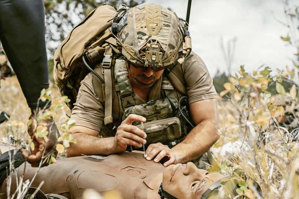 A military medic in full combat gear is performing a medical procedure on a training mannequin in an outdoor field environment. The soldier is wearing multicam camouflage, a tactical helmet with communications equipment, and a plate carrier vest with a large rucksack. This appears to be a tactical combat casualty care (TCCC) training exercise.