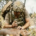 A military medic in full combat gear is performing a medical procedure on a training mannequin in an outdoor field environment. The soldier is wearing multicam camouflage, a tactical helmet with communications equipment, and a plate carrier vest with a large rucksack. This appears to be a tactical combat casualty care (TCCC) training exercise.