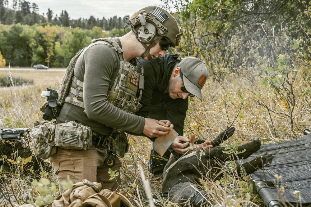 Two individuals are crouched in a field with dry grass and shrubs, appearing to work on something together at ground level. The person on the left is wearing full tactical gear including a helmet, plate carrier vest with an 'EOD' patch, and multicam accessories. The scene appears to depict a tactical training or field exercise scenario.