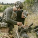 Two individuals are crouched in a field with dry grass and shrubs, appearing to work on something together at ground level. The person on the left is wearing full tactical gear including a helmet, plate carrier vest with an 'EOD' patch, and multicam accessories. The scene appears to depict a tactical training or field exercise scenario.