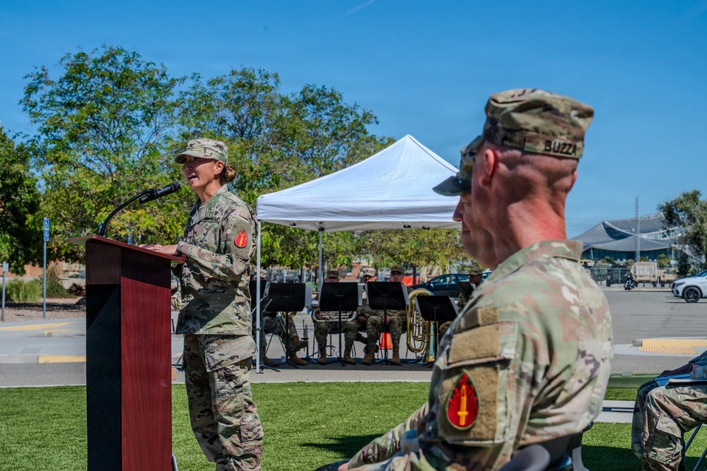 A military ceremony is taking place outdoors on a sunny day, with a female soldier in OCP (Operational Camouflage Pattern) uniform speaking at a wooden podium equipped with a microphone. In the foreground, a male soldier with the name tape 'BUZZA' and a red unit patch is seated or standing with his back to the camera. A military band is visible in the background under a white canopy tent.