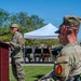 A military ceremony is taking place outdoors on a sunny day, with a female soldier in OCP (Operational Camouflage Pattern) uniform speaking at a wooden podium equipped with a microphone. In the foreground, a male soldier with the name tape 'BUZZA' and a red unit patch is seated or standing with his back to the camera. A military band is visible in the background under a white canopy tent.