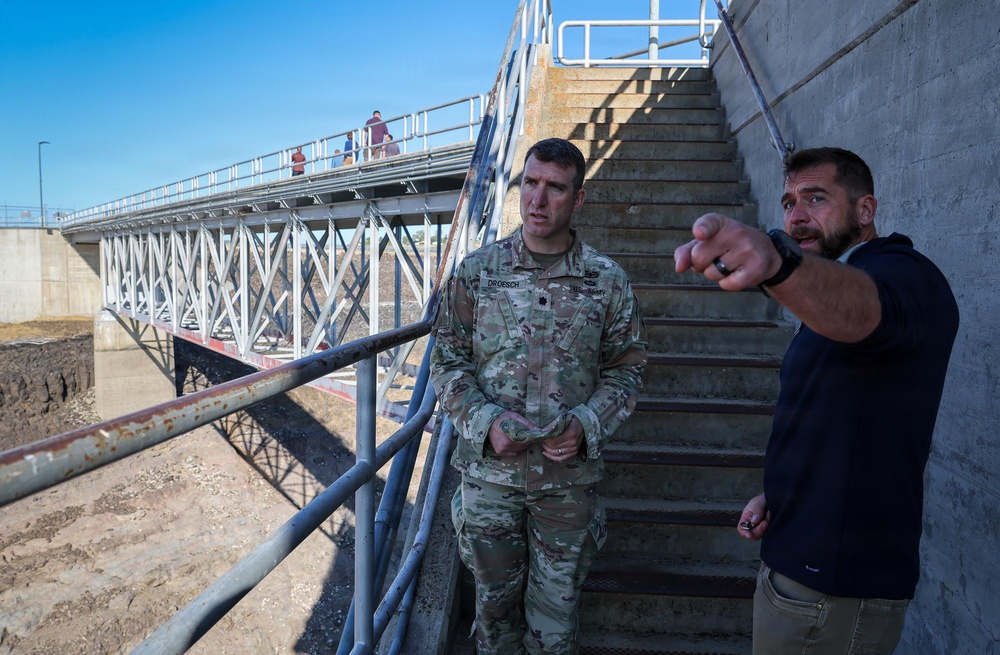Two men stand at the base of concrete stairs near a metal bridge structure at what appears to be a dam, lock, or flood control infrastructure site. A man in U.S. Army camouflage uniform with a rank insignia (Lieutenant Colonel oak leaf) listens while a civilian in a dark hoodie points toward something off-frame, apparently briefing or explaining site details. Additional personnel are visible on the elevated walkway/bridge in the background.