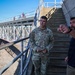 Two men stand at the base of concrete stairs near a metal bridge structure at what appears to be a dam, lock, or flood control infrastructure site. A man in U.S. Army camouflage uniform with a rank insignia (Lieutenant Colonel oak leaf) listens while a civilian in a dark hoodie points toward something off-frame, apparently briefing or explaining site details. Additional personnel are visible on the elevated walkway/bridge in the background.