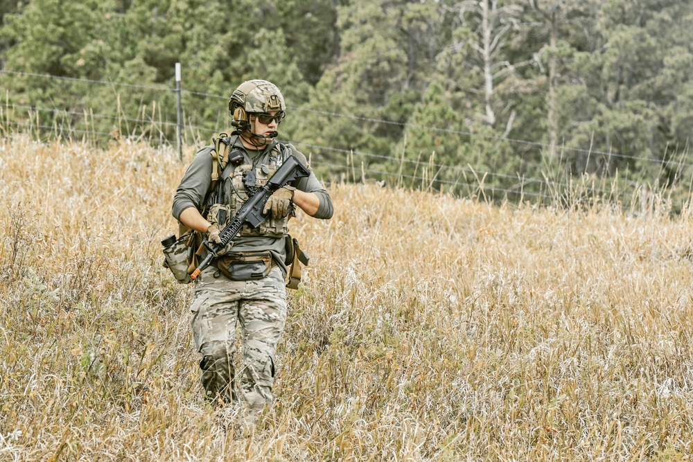 A fully equipped soldier or tactical operator moves through a dry grassland field while checking their watch. The individual is wearing multicam camouflage and carrying full combat kit including a rifle. The setting appears to be a training exercise or field operation in a rural, wooded area.