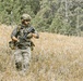 A fully equipped soldier or tactical operator moves through a dry grassland field while checking their watch. The individual is wearing multicam camouflage and carrying full combat kit including a rifle. The setting appears to be a training exercise or field operation in a rural, wooded area.