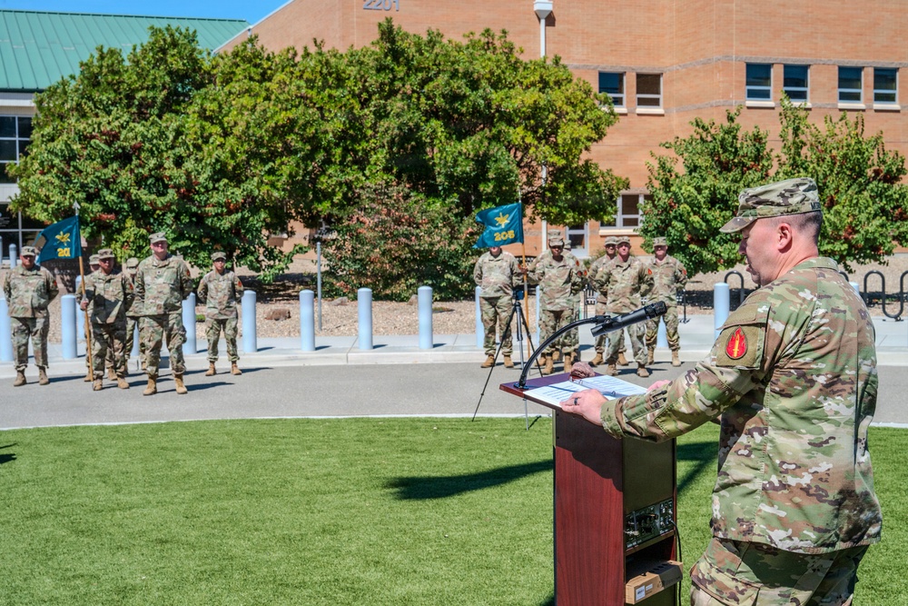 A U.S. Army military ceremony is taking place outdoors in front of a brick building numbered 2201. A soldier in OCP (Operational Camouflage Pattern) uniform is speaking at a wooden podium while a formation of soldiers stands at attention in the background. Two teal/green unit guidon flags are visible among the formation, indicating a formal unit ceremony such as a change of command or activation.