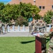 A U.S. Army military ceremony is taking place outdoors in front of a brick building numbered 2201. A soldier in OCP (Operational Camouflage Pattern) uniform is speaking at a wooden podium while a formation of soldiers stands at attention in the background. Two teal/green unit guidon flags are visible among the formation, indicating a formal unit ceremony such as a change of command or activation.