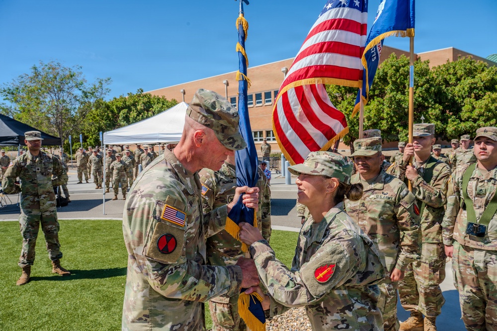 A U.S. Army military ceremony is taking place outdoors, likely a change of command or activation ceremony. Two senior soldiers are exchanging or passing a unit guidon (flag), which is the central symbolic act of such ceremonies. Multiple uniformed soldiers are standing in formation in the background.