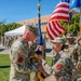 A U.S. Army military ceremony is taking place outdoors, likely a change of command or activation ceremony. Two senior soldiers are exchanging or passing a unit guidon (flag), which is the central symbolic act of such ceremonies. Multiple uniformed soldiers are standing in formation in the background.