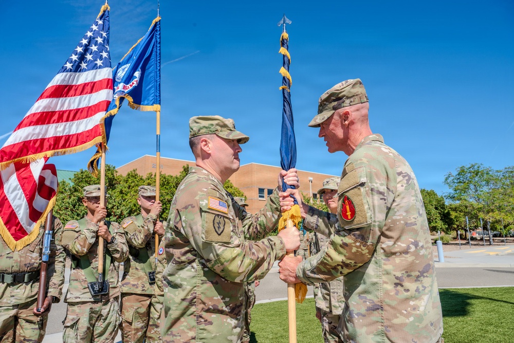 A U.S. Army change of command ceremony is taking place outdoors on a sunny day. Two senior officers are exchanging a unit guidon (flag), which is the central symbolic act of a change of command. A color guard stands in the background holding the American flag and the U.S. Army flag.