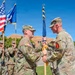 A U.S. Army change of command ceremony is taking place outdoors on a sunny day. Two senior officers are exchanging a unit guidon (flag), which is the central symbolic act of a change of command. A color guard stands in the background holding the American flag and the U.S. Army flag.