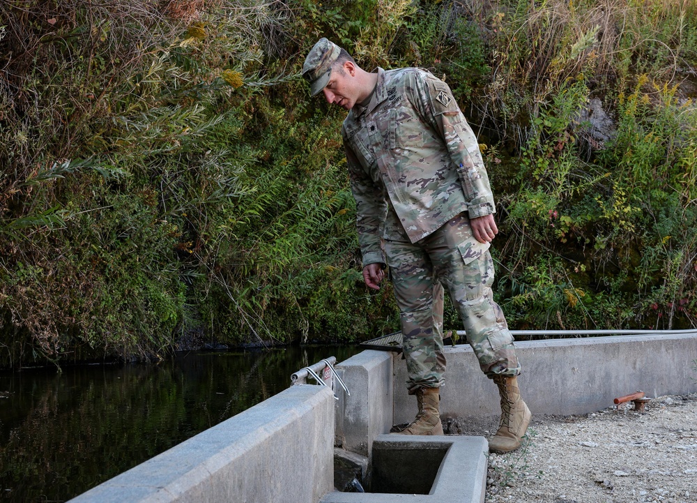 A U.S. Army soldier in OCP (Operational Camouflage Pattern) uniform is inspecting a concrete water control structure near a canal or irrigation channel. The soldier is looking downward at the structure, appearing to assess or examine the water flow mechanism. The setting is an outdoor area with dense vegetation and a water channel in the background.