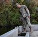 A U.S. Army soldier in OCP (Operational Camouflage Pattern) uniform is inspecting a concrete water control structure near a canal or irrigation channel. The soldier is looking downward at the structure, appearing to assess or examine the water flow mechanism. The setting is an outdoor area with dense vegetation and a water channel in the background.
