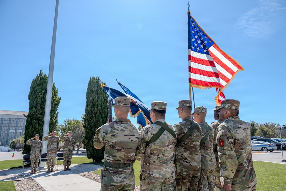 A military color guard ceremony is taking place outdoors on a sunny day, with several soldiers in OCP (Operational Camouflage Pattern) uniforms carrying flags including the United States flag and what appear to be unit or service flags. Two soldiers in the background are rendering a salute toward the color guard. The setting appears to be a military installation with a flagpole, manicured grounds, and institutional buildings visible.