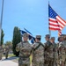 A military color guard ceremony is taking place outdoors on a sunny day, with several soldiers in OCP (Operational Camouflage Pattern) uniforms carrying flags including the United States flag and what appear to be unit or service flags. Two soldiers in the background are rendering a salute toward the color guard. The setting appears to be a military installation with a flagpole, manicured grounds, and institutional buildings visible.