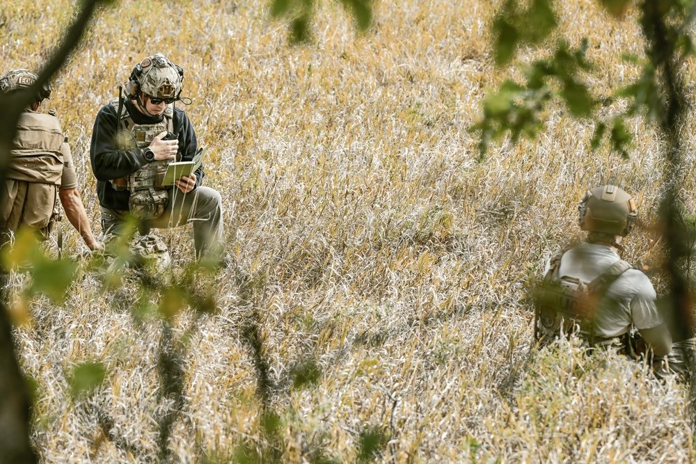 Three individuals in military-style tactical gear are positioned in an open field of dry grass, partially obscured by foreground foliage. The central figure is crouching and examining what appears to be a tablet or document, while a second figure sits to the right and a third is partially visible on the left. The setting and equipment suggest a field exercise, reconnaissance, or operational planning activity.