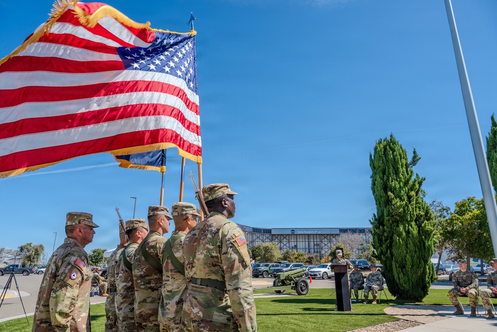 A U.S. Army color guard ceremony is taking place outdoors on a sunny day, with soldiers in OCP (Operational Camouflage Pattern) uniforms carrying the American flag and additional military colors. A speaker stands at a podium in the background while other soldiers observe the ceremony seated and standing. The setting appears to be a military installation or government facility with a large building visible in the background.