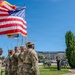 A U.S. Army color guard ceremony is taking place outdoors on a sunny day, with soldiers in OCP (Operational Camouflage Pattern) uniforms carrying the American flag and additional military colors. A speaker stands at a podium in the background while other soldiers observe the ceremony seated and standing. The setting appears to be a military installation or government facility with a large building visible in the background.