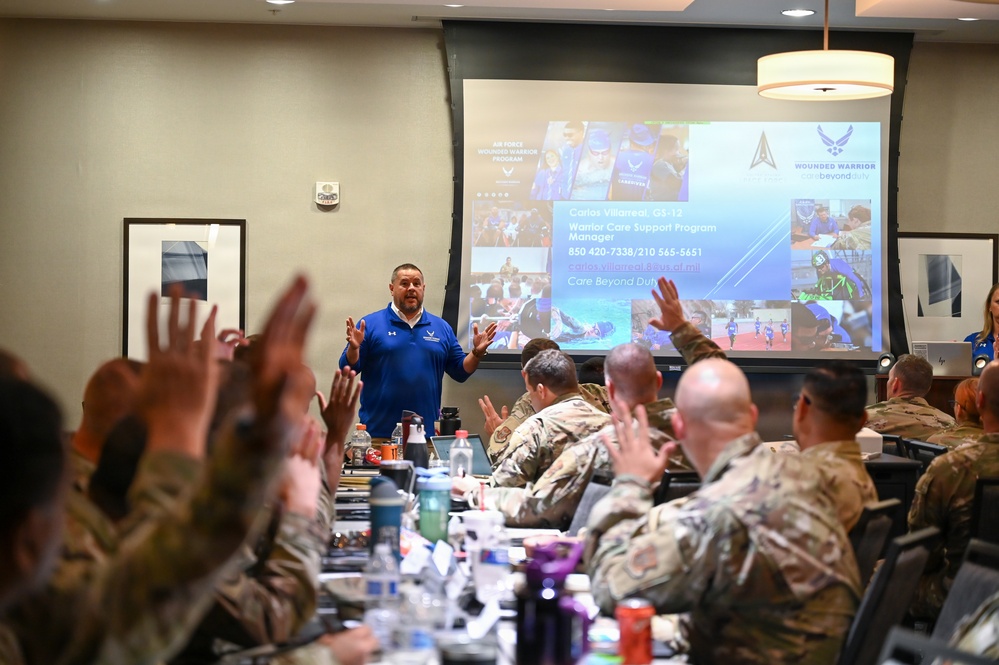 A presenter in a blue polo shirt stands before a group of military personnel in camouflage uniforms during a briefing on the Air Force Wounded Warrior Program. The slide behind him identifies the speaker as Carlos Villarreal, GS-12, Warrior Care Support Program Manager. Multiple audience members have raised their hands, indicating active engagement with the presentation.
