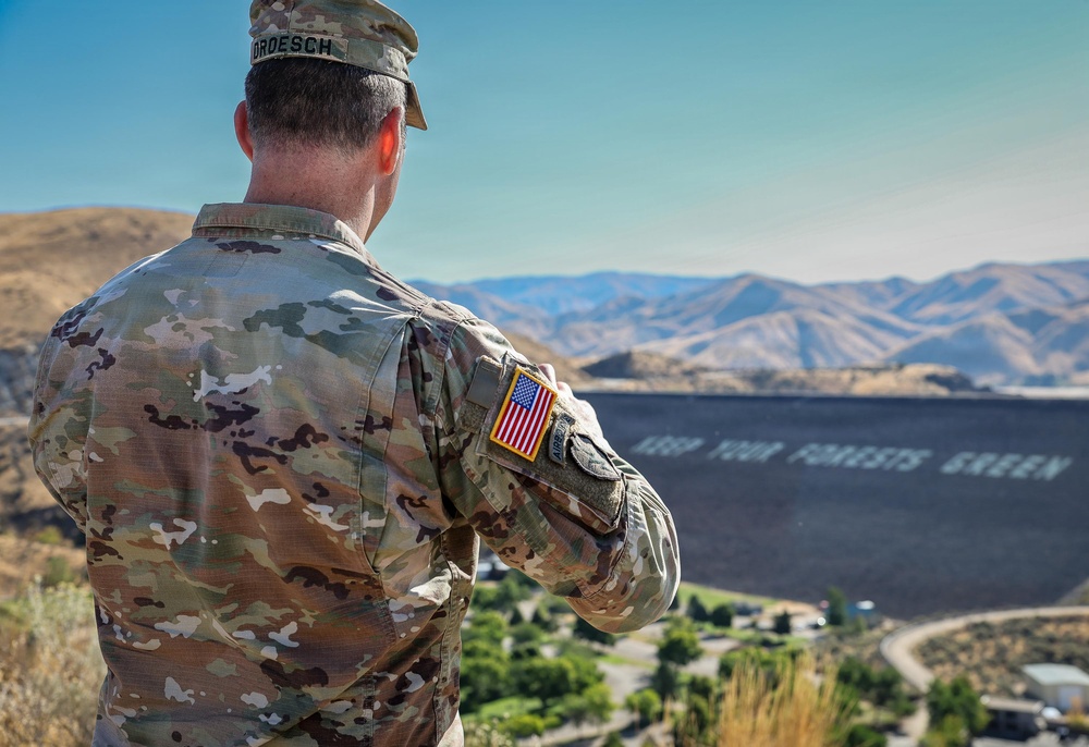 A U.S. Army soldier in OCP (Operational Camouflage Pattern) uniform stands with arms crossed, viewed from behind, overlooking a large dam structure in a mountainous, arid landscape. The soldier's name tape reads 'DROESCH' and an American flag patch is visible on the left sleeve. A message reading 'KEEP YOUR FORESTS GREEN' is painted on the face of the dam.