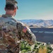 A U.S. Army soldier in OCP (Operational Camouflage Pattern) uniform stands with arms crossed, viewed from behind, overlooking a large dam structure in a mountainous, arid landscape. The soldier's name tape reads 'DROESCH' and an American flag patch is visible on the left sleeve. A message reading 'KEEP YOUR FORESTS GREEN' is painted on the face of the dam.